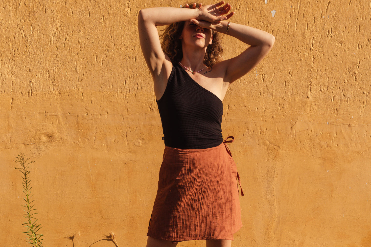 A woman posing outdoors against a textured ochre-colored wall, wearing a black one-shoulder top and a rust-orange textured wrap skirt.